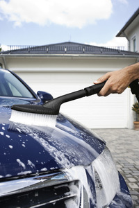 Hand using Kärcher brush to clean a blue car bonnet with soap suds, outdoors in front of a garage.