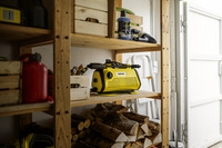 Yellow Kärcher high-pressure cleaner on wooden shelf in garage, surrounded by cleaning supplies and stacked firewood.