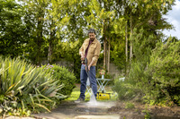 Man using Kärcher high-pressure cleaner on garden path, surrounded by lush greenery and outdoor furniture.