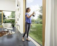Woman cleaning large glass door with Kärcher window vac in modern living room, featuring a chair, wall art, and garden view outside.
