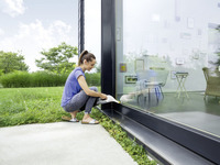 Woman cleaning large glass door with Kärcher window vac, outdoors, grassy area, interior visible through glass with furniture and wall art.