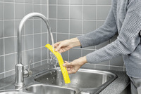 Hands rinsing a yellow Kärcher window vac blade under a kitchen tap, with grey tiled wall background.