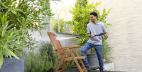 Person using Kärcher high-pressure cleaner on wooden chair in outdoor garden setting with plants and stone wall.