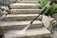 A Kärcher high-pressure cleaner sprays water on outdoor stone steps surrounded by rocks and ferns.