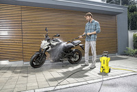 Man cleaning motorcycle with Kärcher high-pressure cleaner outdoors, wooden wall background.