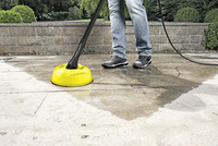 Person cleaning outdoor stone patio with Kärcher surface cleaner, wearing jeans and trainers.