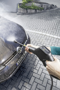 Person using Kärcher high-pressure cleaner on a dirty car bonnet outdoors on a paved driveway.