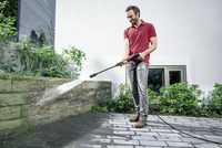 Man using Kärcher high-pressure cleaner on stone wall outdoors, surrounded by greenery and a modern building.