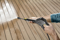 Hands using a Kärcher high-pressure cleaner to spray water on a wooden deck outdoors.