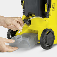Hands connecting a hose to a detergent tank on a Kärcher high-pressure cleaner, with visible wheels and yellow casing.