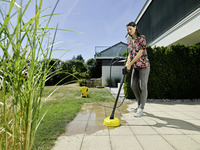 Person cleaning patio with Kärcher surface cleaner outdoors, surrounded by grass and bushes, modern house in background.