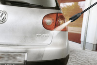 Kärcher high-pressure cleaner spraying water on a dirty Volkswagen car rear, outdoors on a paved surface.