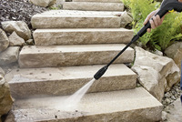 Hand using Kärcher high-pressure cleaner to wash outdoor stone steps surrounded by rocks and ferns.