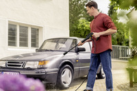 Man using Kärcher high-pressure cleaner to wash a grey car outdoors, near a white building with windows and greenery in the background.