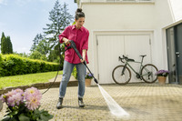 Person using a Kärcher high-pressure cleaner on a patio. A bicycle is parked against a garage door, surrounded by potted plants.