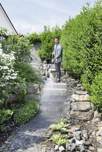 Person using a Kärcher high-pressure cleaner on stone steps in a lush garden with green shrubs and white flowers.