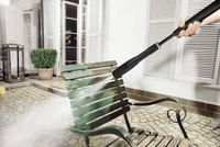 Hand using Kärcher high-pressure cleaner on a green metal chair outdoors, with water spray removing dirt. White shutters and potted plant in background.