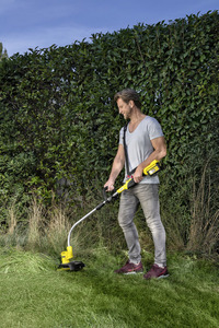 Man using a Kärcher grass trimmer in a garden, standing on a lawn with a tall hedge in the background.