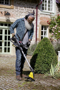 Man using Kärcher weed remover on cobblestone path in garden, wearing gloves and headphones, with a stone house and greenery in the background.