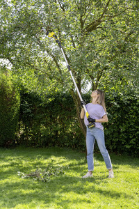 Person using a Kärcher tree pruner to trim branches in a sunny garden, with cut branches on the grass.