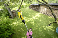 Person using a Kärcher tree pruner to cut a branch in a garden with a woodpile and lush greenery in the background.