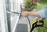 Person using a Kärcher high-pressure cleaner to clean a metal gutter pipe outdoors near a window and garden.