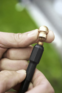 Hand holding a brass nozzle above a black hose connector, outdoors with blurred green background.