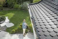 Person using Kärcher high-pressure cleaner to clear leaves from a house gutter, standing on a stone patio in a garden setting.