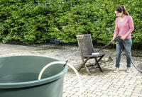 Person using Kärcher high-pressure cleaner on wooden chair outdoors, near a large green water container, with lush green hedge in background.
