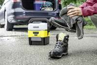 Person cleaning hiking boot with cloth next to Kärcher portable cleaner, car boot open in background, outdoor setting.
