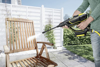 Person using a Kärcher high-pressure cleaner on a wooden chair outdoors, with a white fence and greenery in the background.