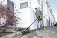 Person cleaning outdoor steps with Kärcher high-pressure cleaner, near a white building and a red-leafed tree.