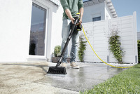 Man cleaning patio with Kärcher high-pressure cleaner, outdoors near white fence and grass.