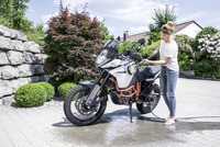 Woman cleaning a motorcycle outdoors with a Kärcher high-pressure cleaner, surrounded by stone wall and lush greenery.