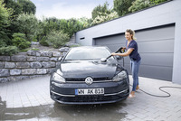 Person cleaning a black Volkswagen car with a Kärcher high-pressure cleaner in a driveway, surrounded by greenery and a stone wall.