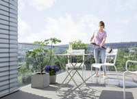 Person cleaning a white chair on a balcony with a Kärcher high-pressure cleaner, surrounded by plants and outdoor furniture.