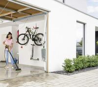 Person cleaning garage floor with Kärcher high-pressure cleaner; bicycle and scooter mounted on wall; outdoor greenery visible.