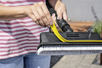 Hands holding a Kärcher scrub brush with white bristles, outdoors. Person wearing a striped shirt and jeans.
