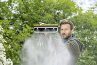 Person using a Kärcher high-pressure cleaner outdoors, surrounded by greenery, with water mist spraying from the device.