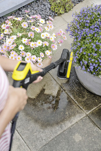 Person cleaning patio with Kärcher high-pressure cleaner near pots of pink and purple flowers.