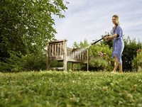 Woman cleaning wooden bench with Kärcher high-pressure cleaner in a garden, surrounded by greenery and flowering plants.