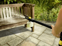 Person using a Kärcher high-pressure cleaner to spray water on a wooden bench in a garden setting.