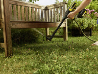 Person using a Kärcher high-pressure cleaner to wash a wooden garden bench, surrounded by grass and flowers.