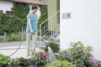 Person watering garden plants with a Kärcher garden hose in front of a house numbered 16, with ivy-covered walls in the background.