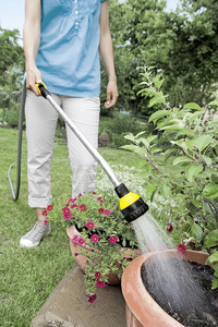 Person watering garden plants with a Kärcher garden hose sprayer, wearing a blue shirt and white trousers outdoors.
