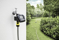 Kärcher water timer attached to outdoor tap, controlling a sprinkler watering a lush green garden.