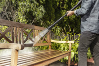 Person cleaning a wooden garden bench with a Kärcher high-pressure cleaner, surrounded by lush greenery.