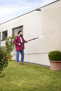 Person using a Kärcher high-pressure cleaner to wash an exterior wall in a garden setting, surrounded by potted plants.
