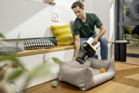 Person using a Kärcher handheld vacuum cleaner on a pet bed in a bright, modern living room with cushions and toys nearby.