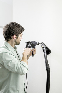 Man using a drill on a white wall, with a Kärcher dust extractor attached to collect debris. Indoor setting.